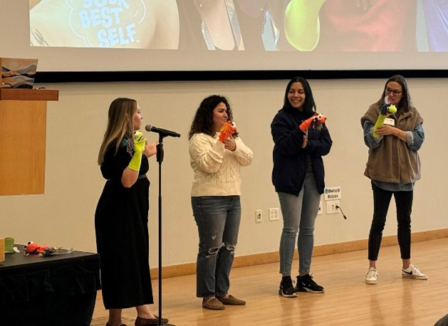 Four women stand in the front of the room, holding sock puppets. One speaks into a mic.