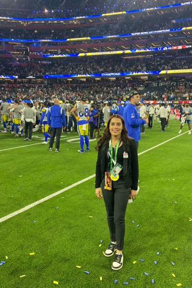 Xaimara Coss stands on a football field with NFL players and staff in the background.