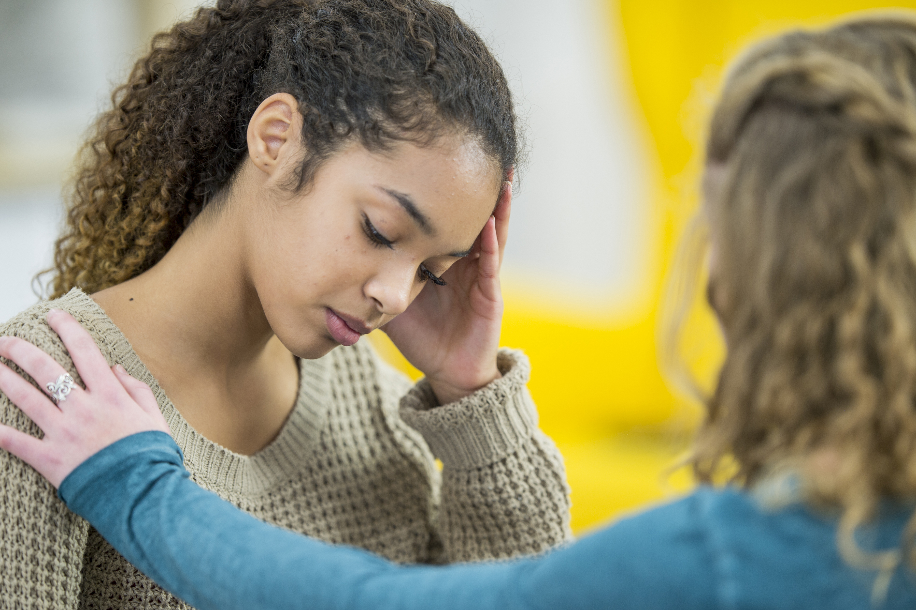 Two young women are indoors in a bedroom. They are wearing casual clothing. One woman looks sad, and she is being comforted by her friend. (Getty Images)