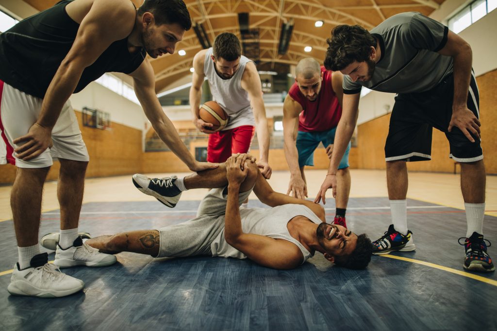 Young African American basketball player experiencing pain while injuring his leg during the match. His teammates are supporting him. (Getty Images)