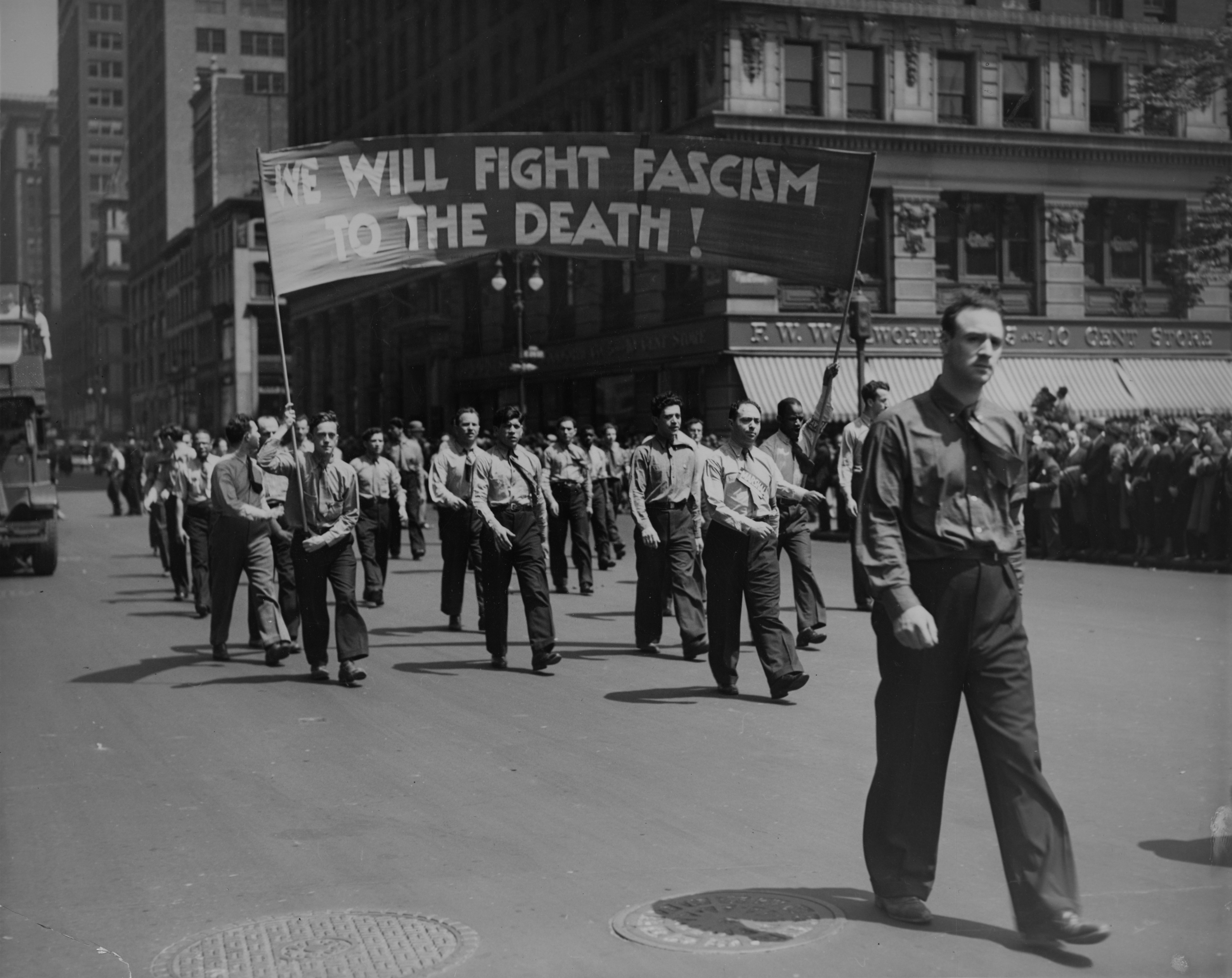 An archival photo from a May Day parade in New York City in the 1930s, with men marching under a banner that says "We Will Fight Fascism to the Death."