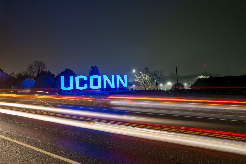 The UConn sign in Storrs, lit up at night, with light steaks from passing traffic