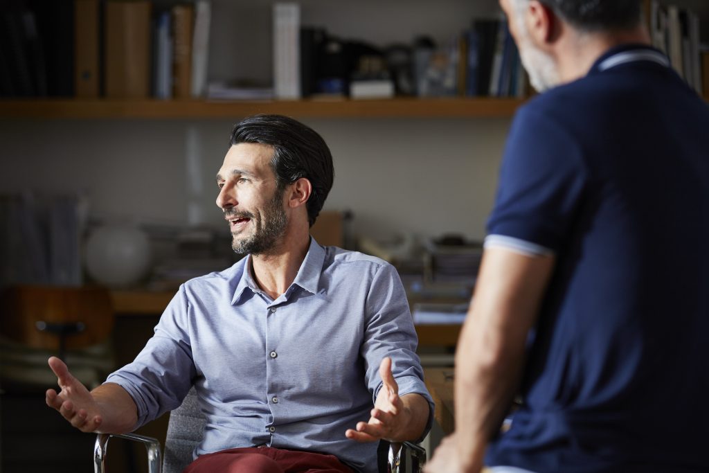 A seated man gesturing with both hands while speaking.