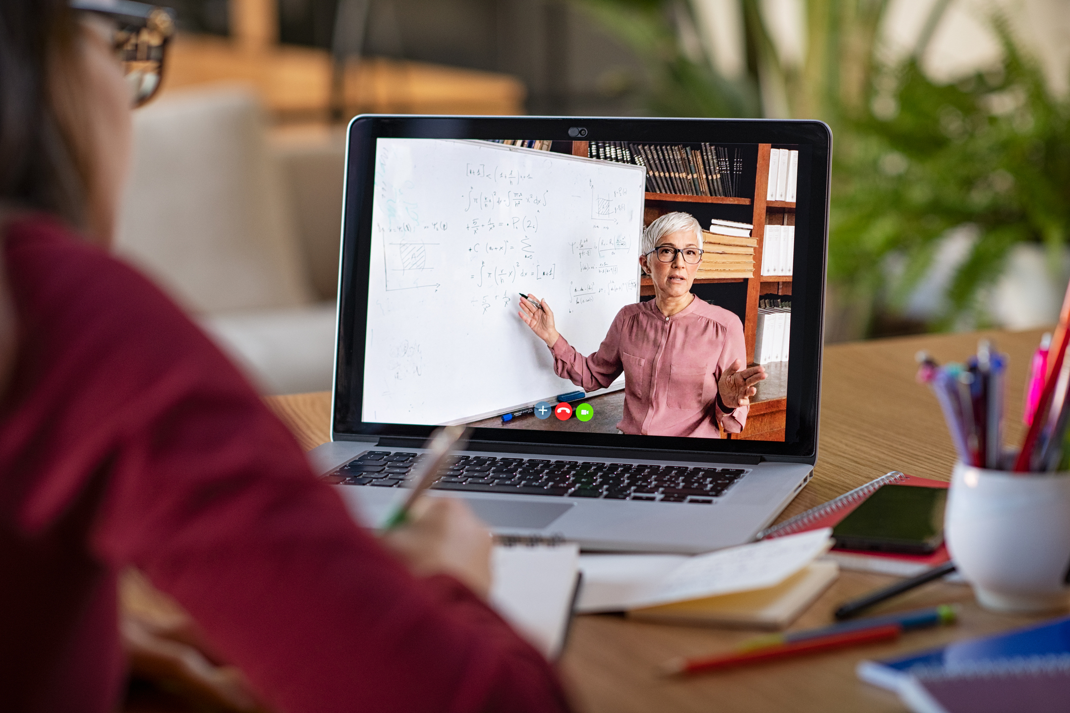 Young student watching lesson online and studying from home. Young woman taking notes while looking at computer screen following professor doing math on video call. Latin girl student studying from home and watching teacher explaining math formula on video chat.