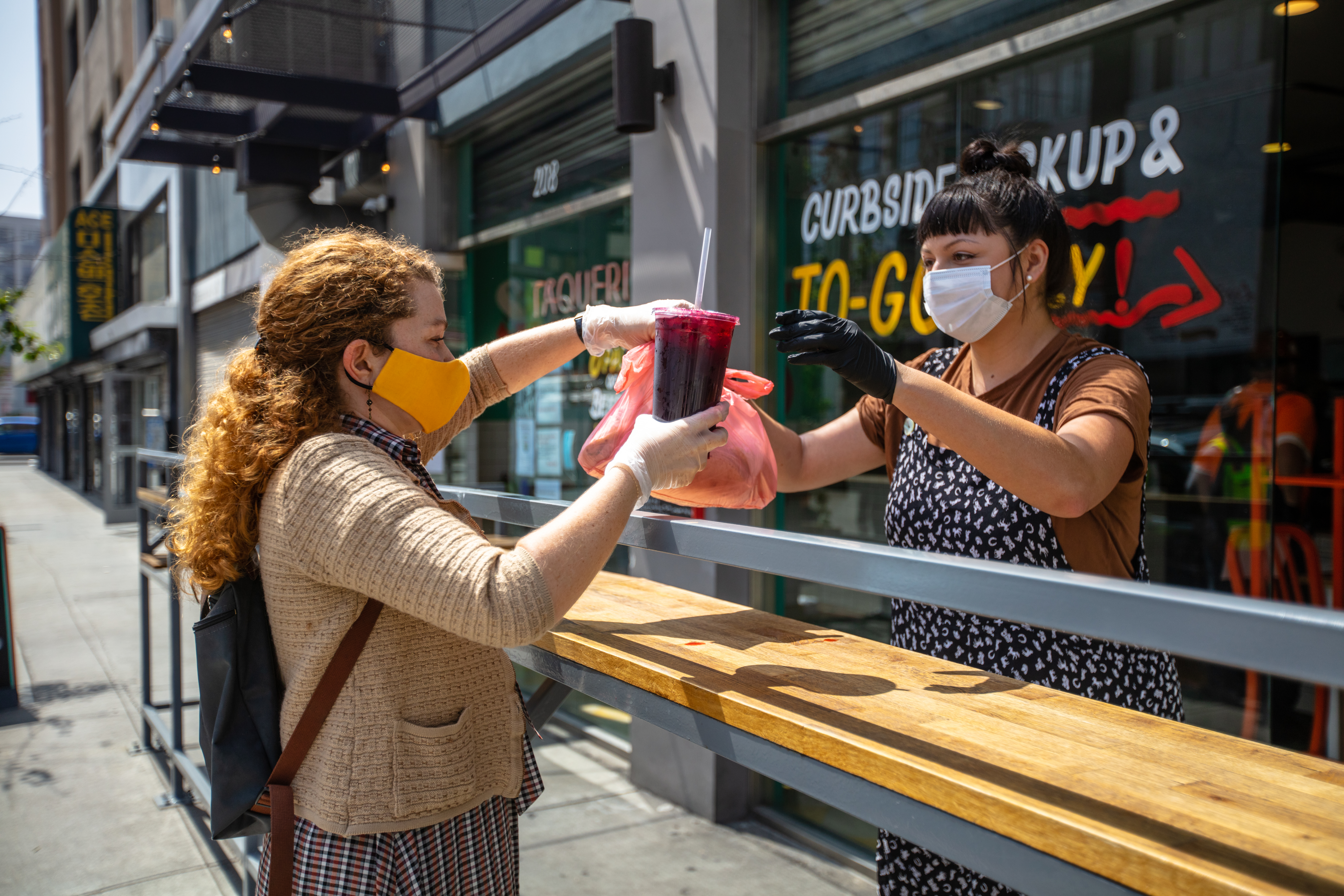 A Mexican restaurant adapts to the Covid-19 lockdown. The owner hands an order to a customer outside the restaurant; they are both wearing gloves and masks.