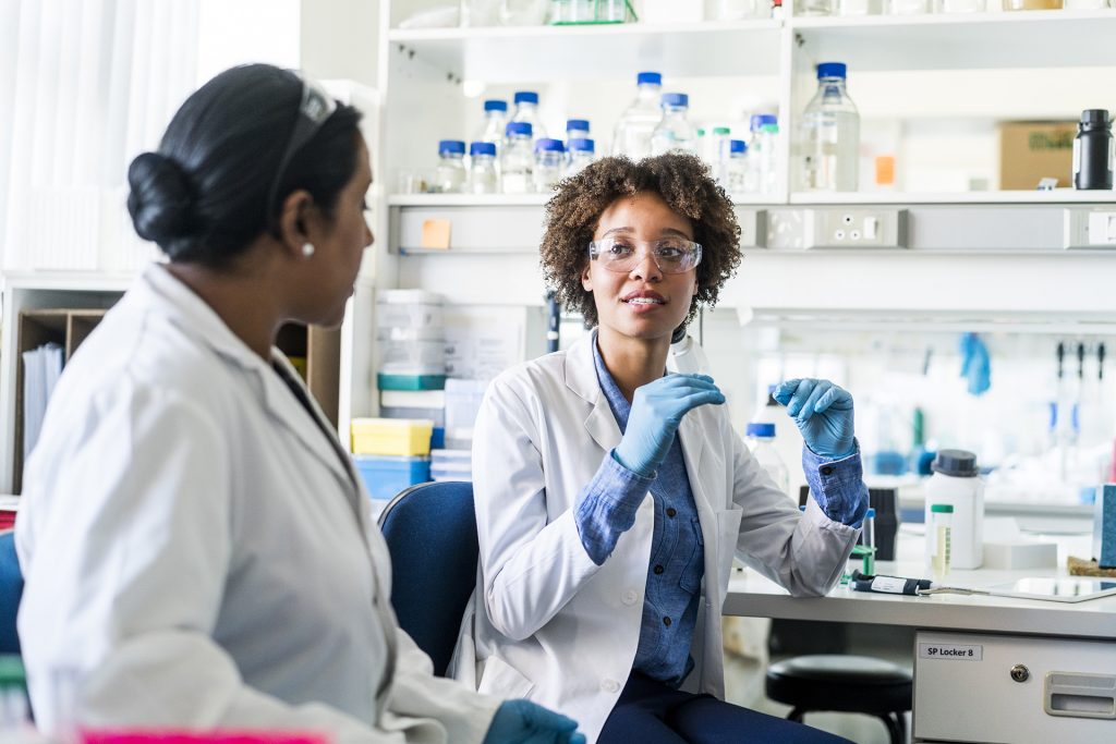 Young scientist discussing with colleague. Female researcher is sharing her ideas during meeting. They are wearing uniforms in laboratory.