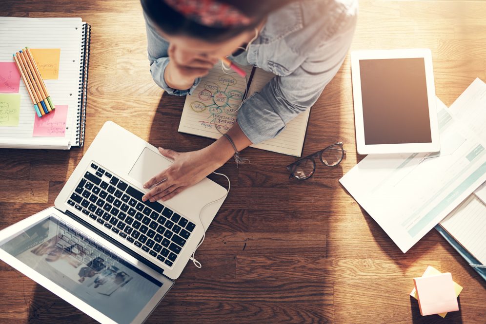 A young entrepreneur works on her laptop, with papers and a tablet computer on the table beside her.