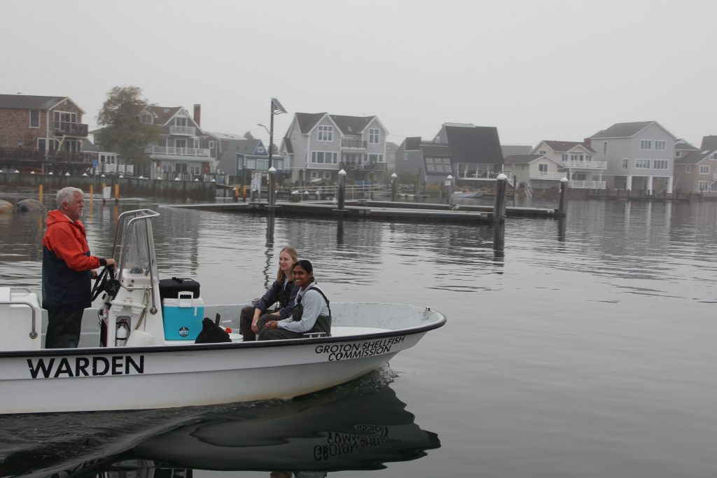 Ted Rathjen, senior shellfish warden for Groton, steers the boat as Kaitlyn Campbell, post doctoral student in the Brandt Lab at UConn, and Anika Agrawal, doctoral student in the lab, prepare to collect oysters and mussels to test for PFAS in shellfish tissue in an earlier project led by Christopher Perkins, who was chosen for one of the CECs project research awards.