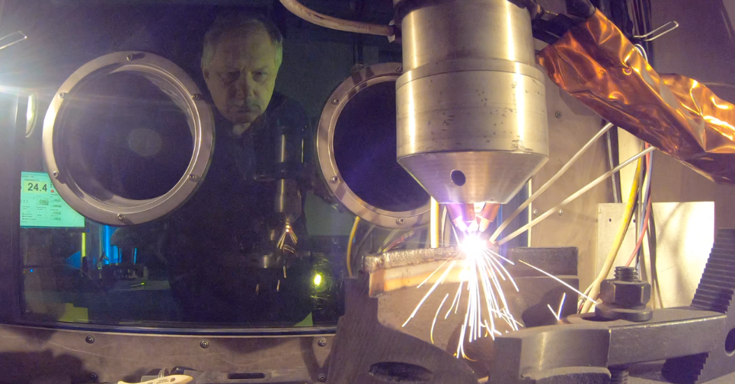 In the background, a man stands behind a sheet of protective glass and monitors the operations of a machine. In the foreground, the machine sends off a cascade of sparks.