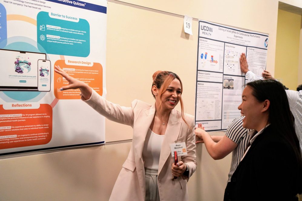 A young person with pink hair, wearing a pink blazer, smiles as she gestures to a poster behind her. Another student smiles at her.