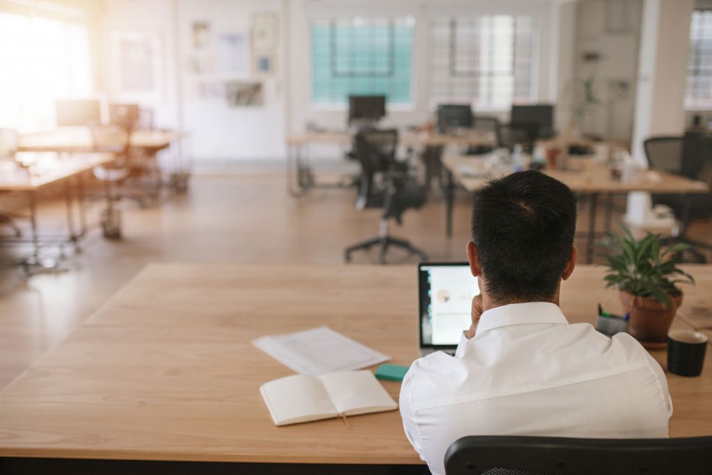 Rearview of a businessman sitting at his desk using a laptop and going over paperwork while working alone in a large modern office