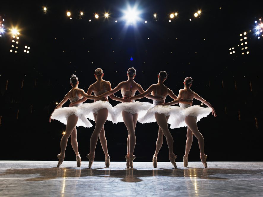 Five ballerinas en pointe on stage, arms around each other, rear view