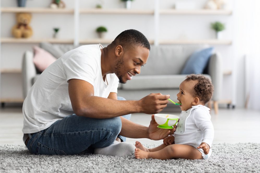 A young father sits on a carpeted floor, spoonfeeding his infant child.