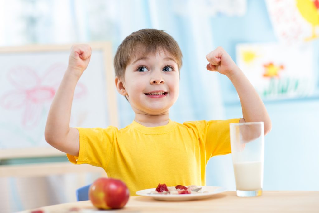 A happy child in a yellow shirt lifts his arms while sitting at a table, with an apple and a glass of milk in front of him.