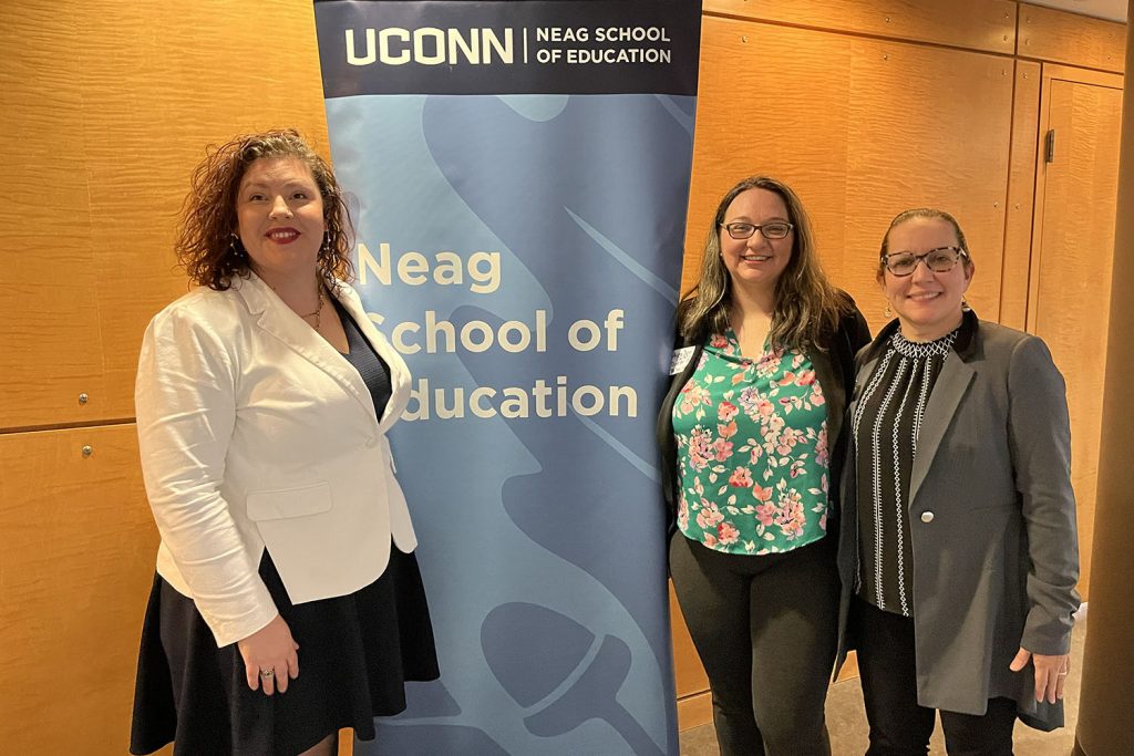 Three female educators stand next to blue Neag School banner.