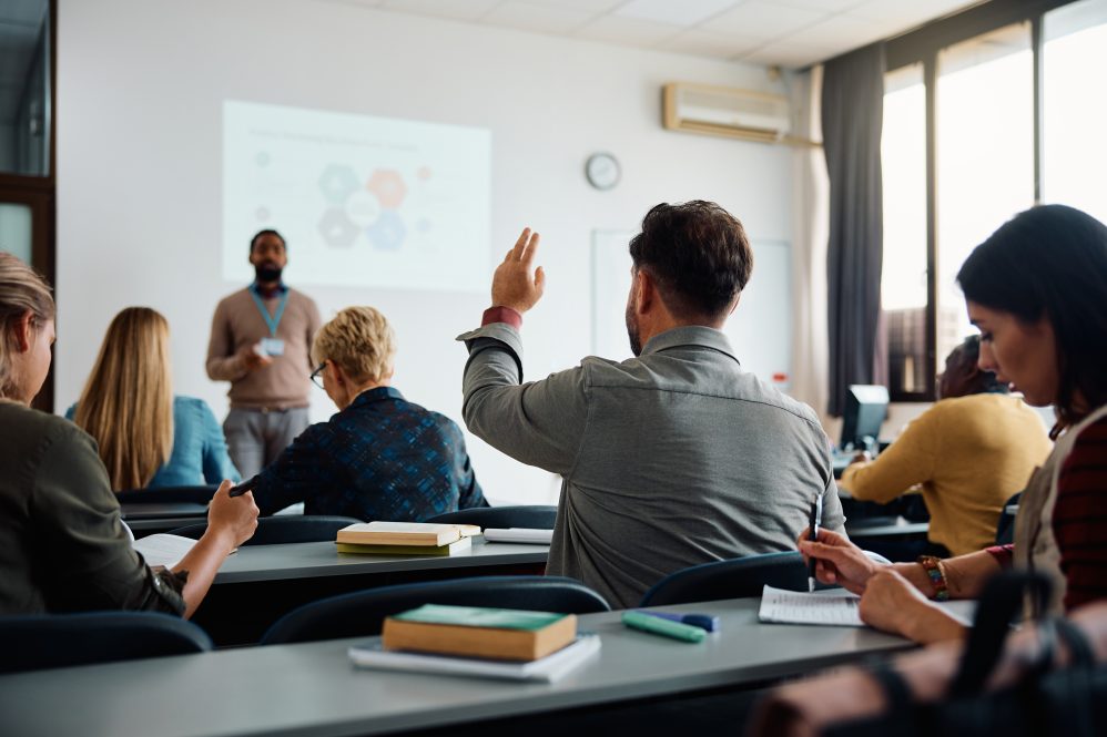 Back view of older student raising his hand to answer teacher's question during class.