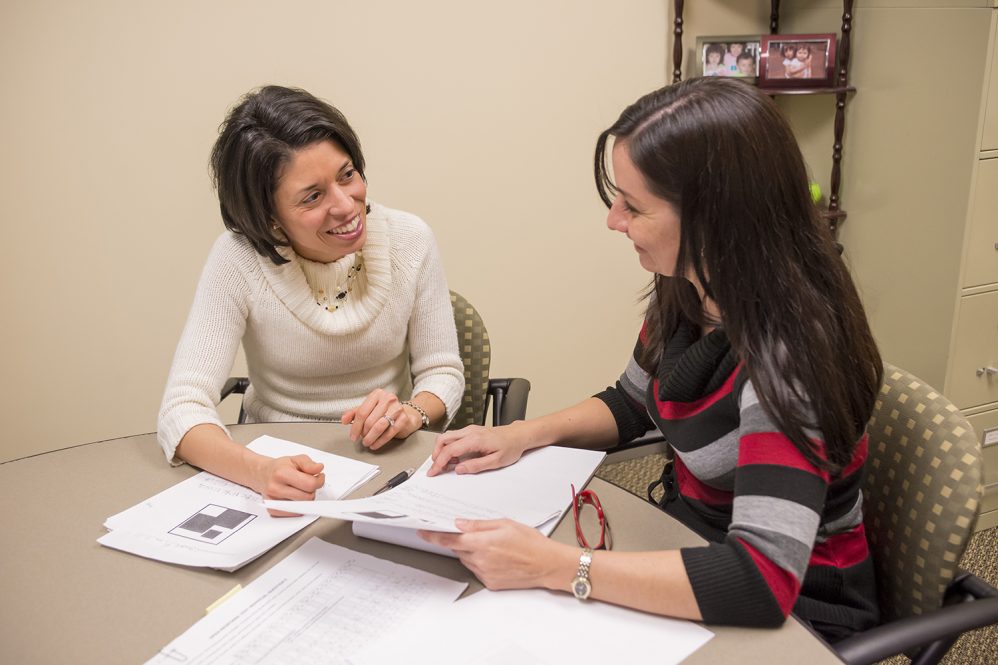 A female professor sits at a table and instructs a female graduate student.