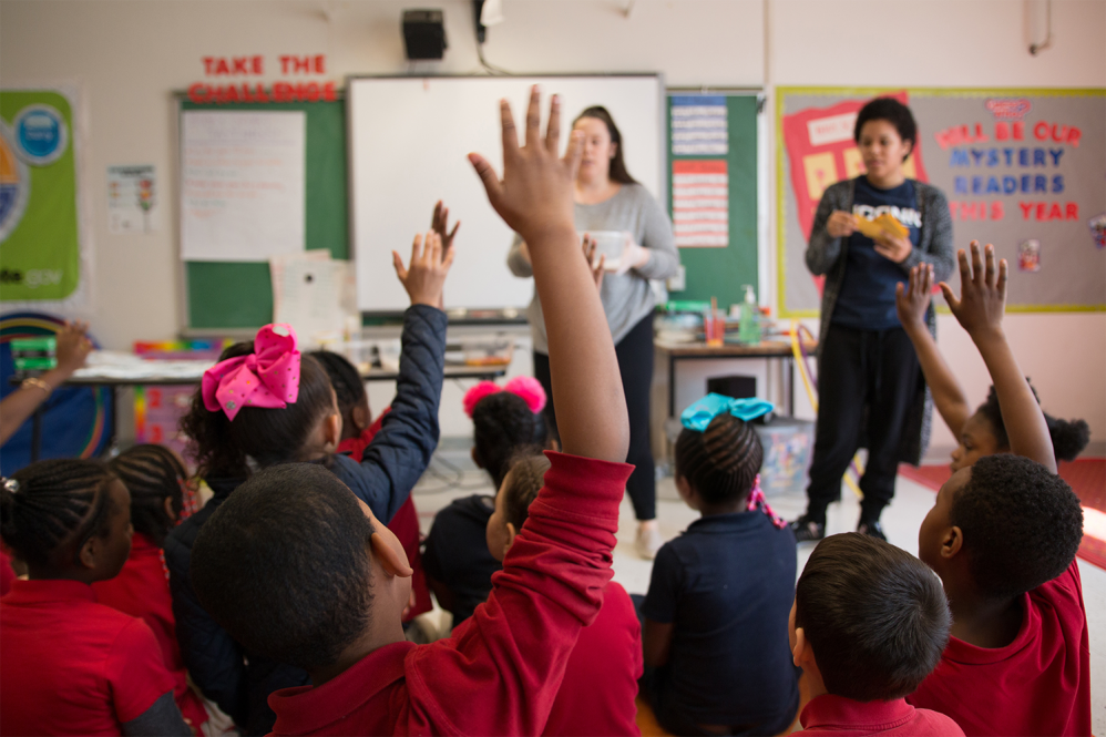 A college student and teacher stand at the front of a classroom, facing a group of elementary students sitting on the floor and raising their hands.