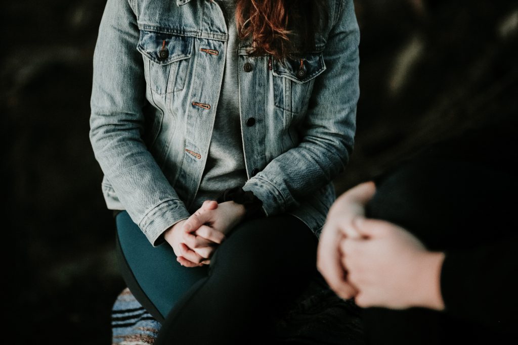 Two people sit near one another, each with their hands clasped together in their lap, as if having a vulnerable conversation.