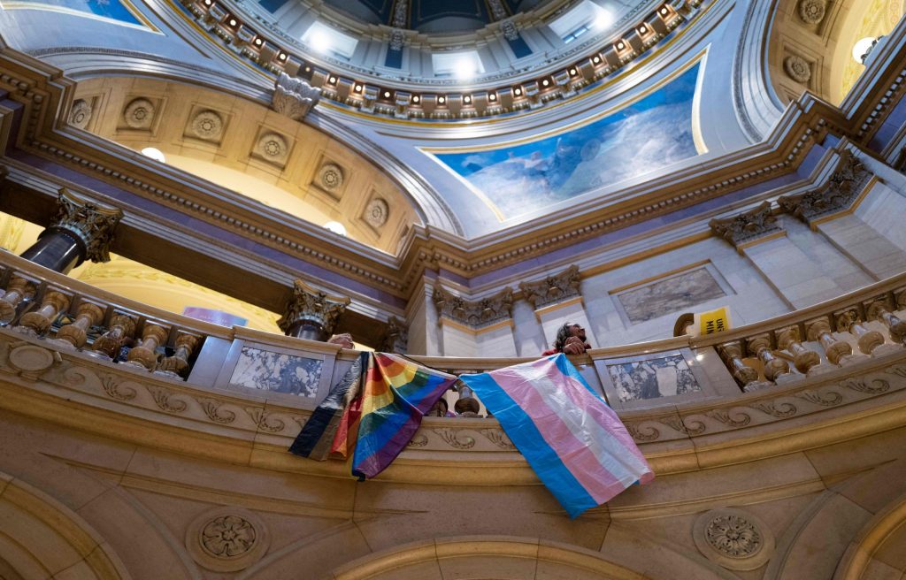 A Pride flag and a trans flag hang side by side over a railing inside a state capitol building.