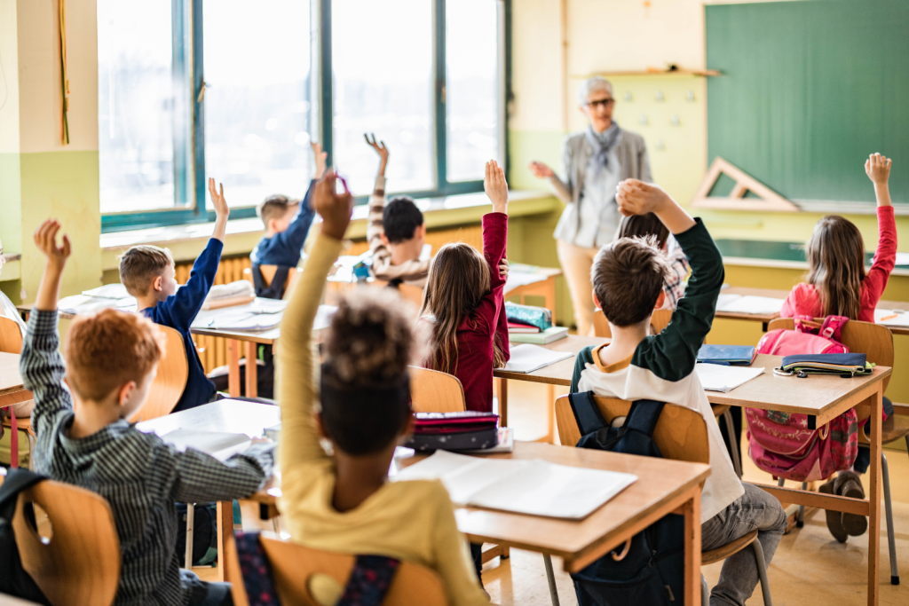 Rear view of elementary students raising hands on a class.