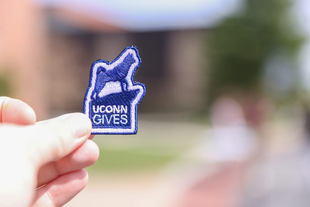 A hand holds up a sticker with the UConn Gives logo.