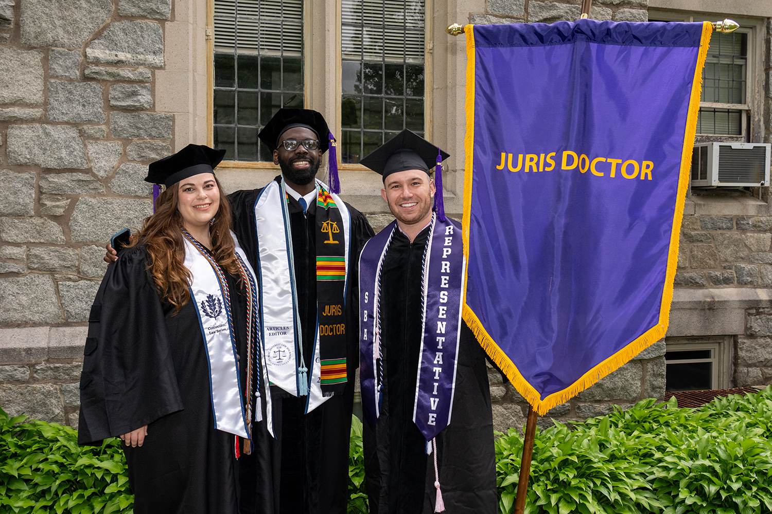 Three UConn law graduates in robes and caps with Juris Doctor flag