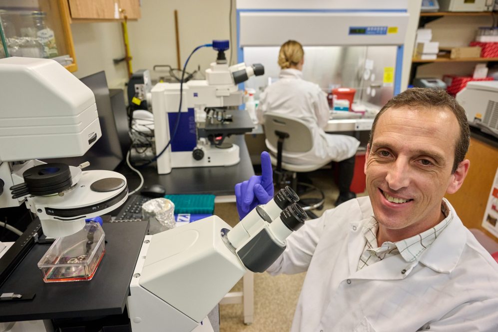 A man in a labcoat smiles in front of a microscope, holding up his index finger.