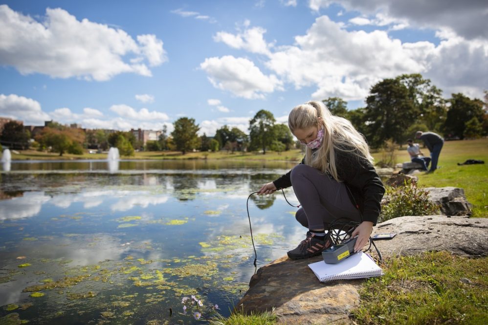 A girl wearing a mask is standing by a lake measuring physiochemical conditions