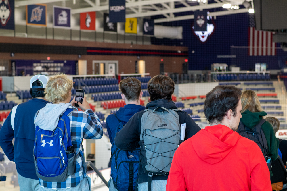 A group of high schoolers stand inside the Toscano Family Ice Forum at UConn Storrs. One student takes a photo with their cellphone.