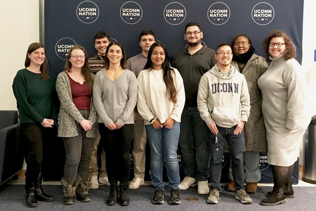 A group of 8 college students and 2 college professors pose in front a navy blue backdrop that reads UConn Nation.